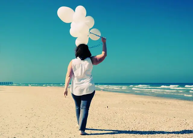 A woman walking on the beach with balloons in her hand.